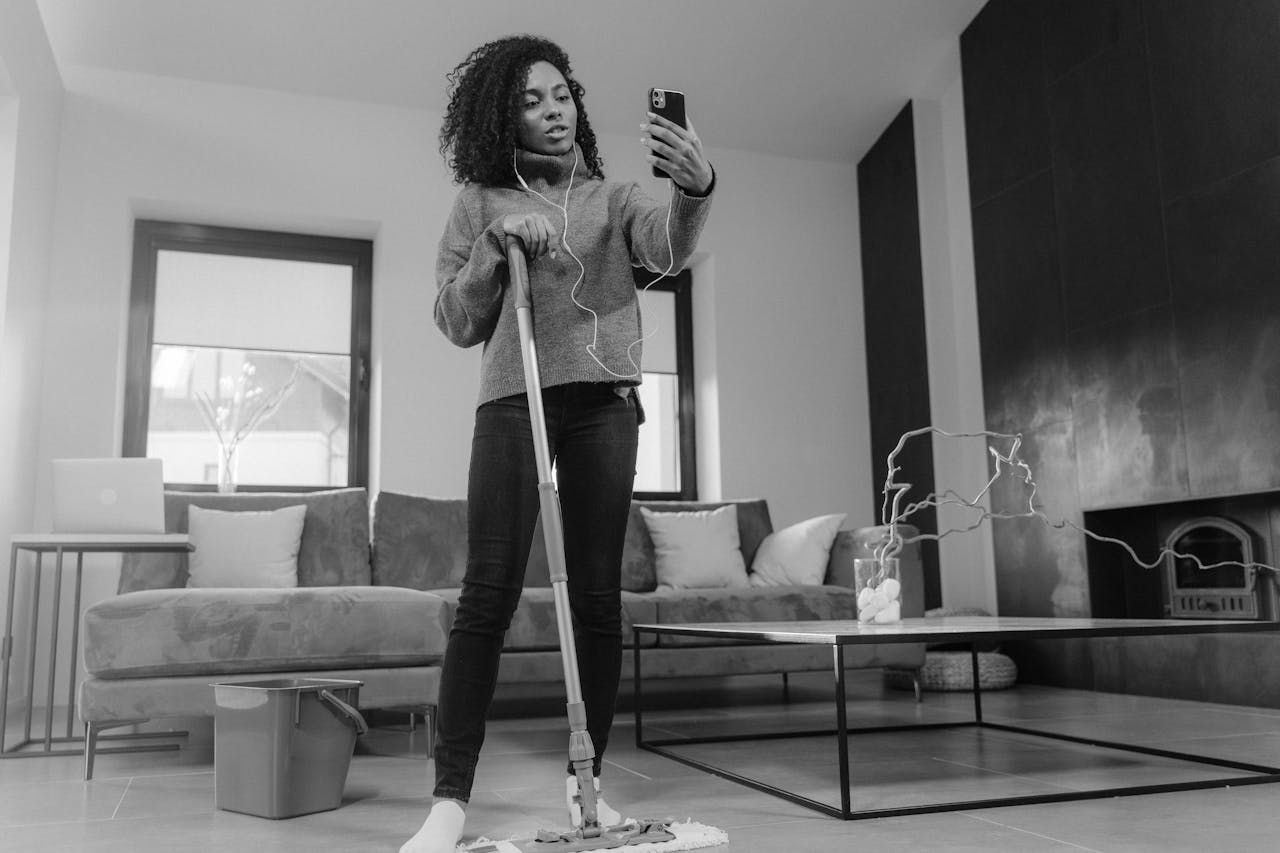 Woman multitasking by mopping the floor and video calling in a modern living room.